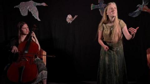 Ailsa Dixon is sitting down on the left playing a cello. She has a bow in her hand and is looking over to Ffion Phillips who is on the right.
Ffion has very long blonde hair. She is wearing a long green dress with no shoes on. 
Ffion is mid performance and holding her left arm up. 