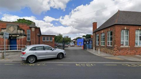 The main entrance to St Peter's C of E Primary School on Cambridge Street it is a single red brick building with slate pitched roofs. A silver car is outside waiting to go through the security barrier