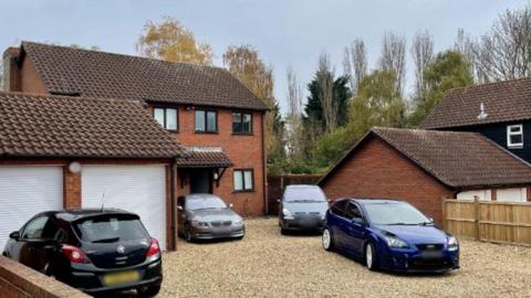 A redbrick house in Holywell Place in Milton Keynes. It is a large detached house with a double garage which has white doors. There are four cars parked on the large gravel driveway in front of the house.