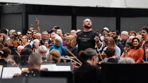 A conductor wearing a black T-shirt that reads "Birmingham forever" leads a large orchestra playing a concert in a station, while a crowd watches behind him.