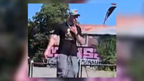 A grainy image of a white man in grey jogging bottoms, black T-shirt and a grey cap. In the background is a building with graffiti on it, surrounded by bushes. 