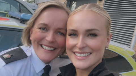 Supt Helen Kenny (left) and her daughter PC Molly Clague (right) both have blonde hair and are in their police uniform smiling at the camera and stood in front of police vehicles.