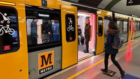 A young woman walks with a backpack walks towards the open doors of the new train. There are a few passengers on the yellow and black carriage which has a sign for Monkseaton. 