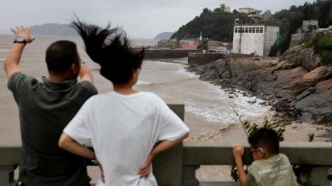 A man, woman and child stand behind a cemented barricade with backs to the camera, looking at muddy waters in a coastal region. There is a rocky shore and a few buildings to the right. The woman's hair is tossed into the air and the child is holding on to the barricade.