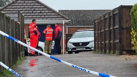A view down a Tarmac driveway, with wooden fencing either side. At the end of the driveway five figures stand close together. One man is wearing a hi-vis orange jacket with FIRE printed across the back and khaki trousers, another is in hi-vis orange overalls. A police car is parked to the right of the group. Blue and white police tape cuts diagonally across the foreground. The ground is wet from rain.