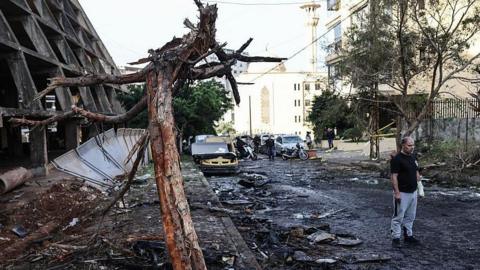 A man stands in the street next to a building and tree which are both heavily damaged and blackened. Rubble lines the street and cars in the background are also damaged.