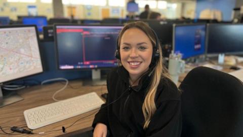 Rhiannon Gardener, wearing a black fleece and headset, sits at a desk in the police control room