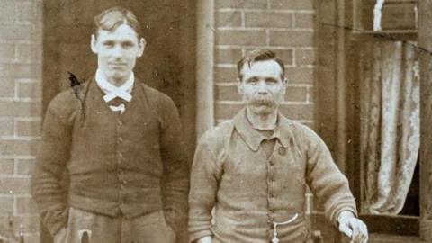 A young man, with a middle parting hair cut and wearing a cardigan, is standing next to his father who has a very thick moustache. He is wearing a jacket, with a pocket watch extending from it. 