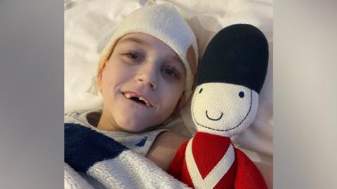 Toby laying in bed next to a knitted toy soldier. He has a bandage on his head and is wearing a grey vest. He is smiling at the camera.