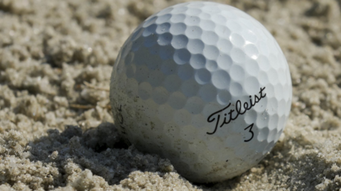 A golf ball in some sand. "Titleist 3" is written on the dimpled white ball.
