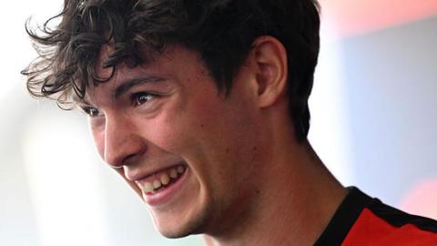 Haas racing driver Ollie Bearman smiling at the camera. He's facing to the side so you can see the profile of his face. He has brown eyes and brown curly hair. He's wearing a red t-shirt with a black collar