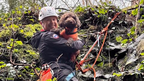 A mountain rescue volunteer strapped with ropes smiles at the camera while holding the dog.