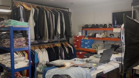 A warehouse lined with clothing rails and shelves full of clothing and trainers. In the centre of a room, there is a table with a laptop and bagged items on it. 