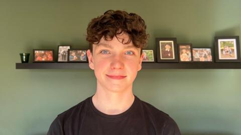 Fiachra, air a bheil dyslexia, na sheasamh air beulaibh chamera agus lèine dhubh air. 