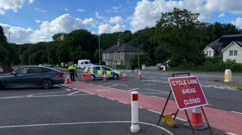 A road closure with police tape and police cars. There is a police officer stood behind some tape attached to traffic cones, along with a small police car next to him.