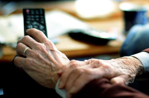 An older person holding a television remote control. The person's hands are in sharp focus, but objects in the room they are sitting in are blurred.