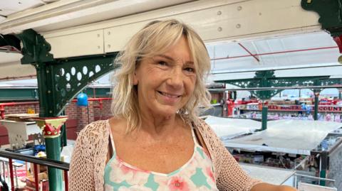 Linzy has shoulder-length bloned hair with a fringe. She is pictured smiling and looking into the camera. She's wearing a blue top with pink flowers and a white knitted cardigan. She's stood on am upstairs mezzanine of an indoor market, which has green wrought iron pillars and a white roof. Beyond Linzy you can see the tops of the market stalls. 