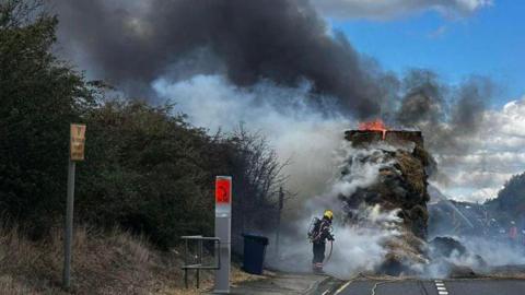 A firefighter in full gear battles a large roadside blaze involving a burning stack of hay or straw on a lorry. Thick black and white smoke billows into the sky as hoses stretch across the road and pavement. The A14 is bordered by trees and bushes, under a partly cloudy daytime sky.