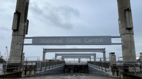 The entrance of the National Glass Centre in Sunderland. A large grey sign is suspended between two high cement pillars. A sloped path leads to the glass doors of the building. The fenced off roof of the building is visible in the distance.