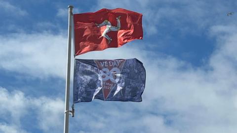 A red Manx flag with a yellow and white triskllion flies on a flag pole. Underneath there is a blue VE Day 80 flag. The sky behind is blue with white clouds.