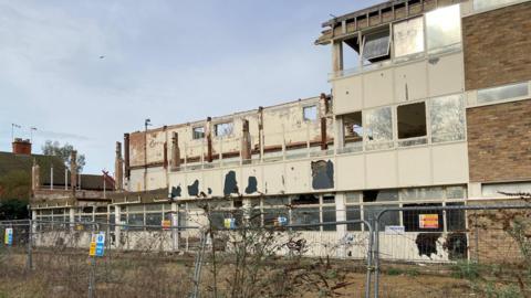 A derelict factory building behind some metal barriers. The roof of the building has collapsed and parts of the exterior walls have fallen down. Several of the remaining windows have been smashed or are missing.