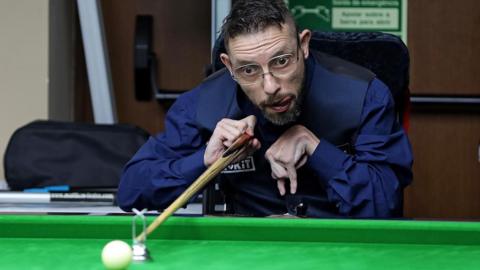 Matt Lester playing snooker. He is using a metal tool on the snooker table to rest the snooker cue on, which helps him to bridge. 