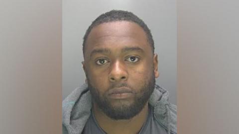 Custody photo of Nabil Aziz against a plain grey background. He has dark hair, brown eyes and a beard. He wears a grey hoodie, grey top and is looking directly at the camera. 