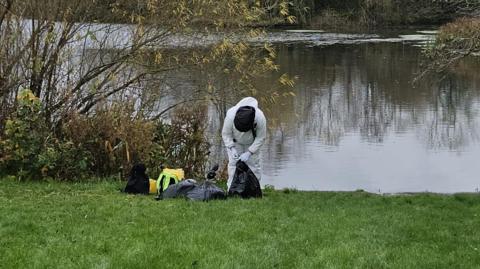 Person wearing white suit stood near lake carrying a black plastic bag. He is stood near to a tree and you cannot see their face.