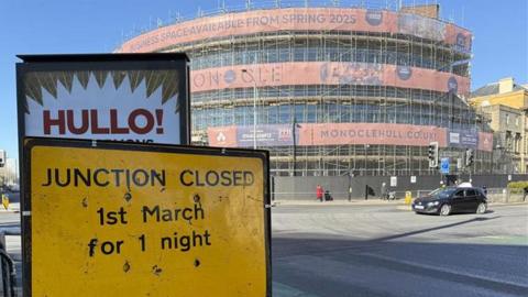 A resurfaced road junction in a city centre. In the foreground is a yellow roadworks sign that reads: "Junction closed 1st March for 1 night". Behind it is a billboard with a poster reading: "Hullo". In the background is a large curved office building which is covered in scaffolding and netting. Banners attached to the scaffolding read: "Monocle", and advertise office space available from spring 2025.