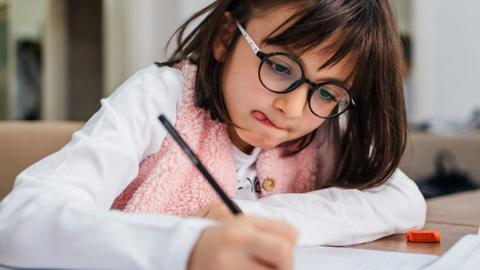 A young girl wearing glasses writing at a table.