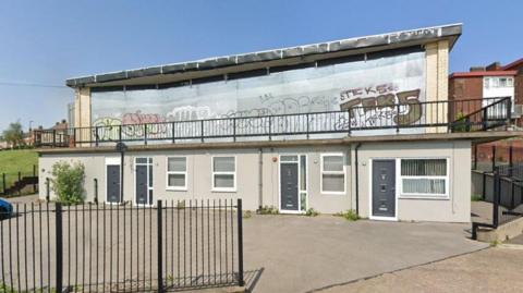 A Google Street View image of the former Byard's Leap pub. The top floor of the two-storey building is boarded up while on the lower level there are four grey doors leading into small offices or homes.