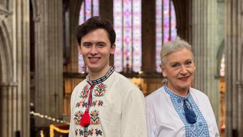 A young man with brown hair is wearing a cream top with colourful embroidery on the front and red tassels. A woman next to him has grey hair and is wearing a white shirt with blue embroidery and tassels. They are standing together in a church with string lights in the backgound