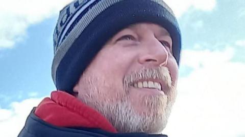 Corporal David Thorne, man smiling, looking away from camera, with a grey beard and wearing a blue and grey wool hat.