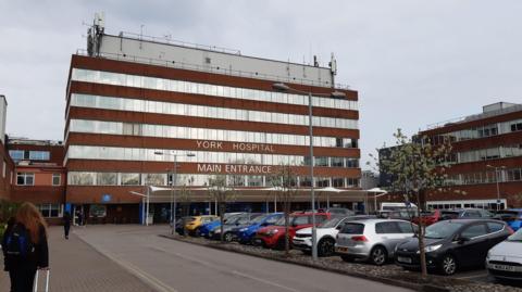 A five-storey red brick building with windows along the width of each floor and the words York Hospital Main Entrance written on it in white lettering. I the foreground is a car park full of cars.