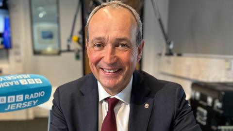 A mid shot of Deputy Lyndon Farnham. He has very little hair and is smiling into the camera. He is wearing a suit, with a maroon tie.