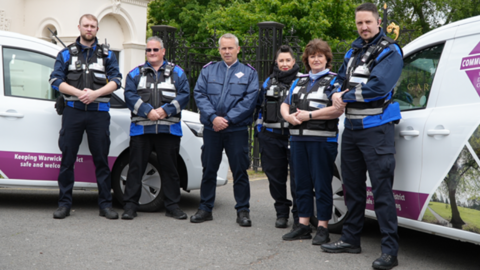 Six people in uniform stand in front of two white vans, looking at the camera. Most have their hands clasped in front of them, and five of the six are wearing tactical vests, with reflective strips and radios attached.