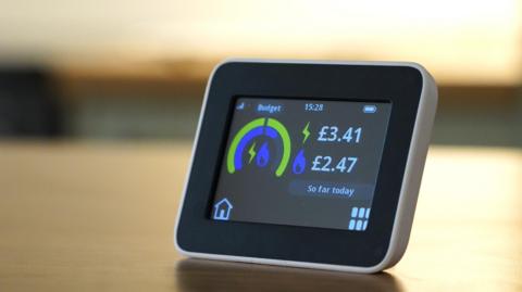 A small smart electricity meter on a table. It has a display that shows a graphic in blue and green contrasting the budget vs the cost of the energy used so far.