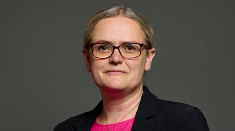 Lizzi Collinge looking directly into the camera. She has blonde hair, parted just off-centre, and is wearing glasses and a black blazer with a pink top.