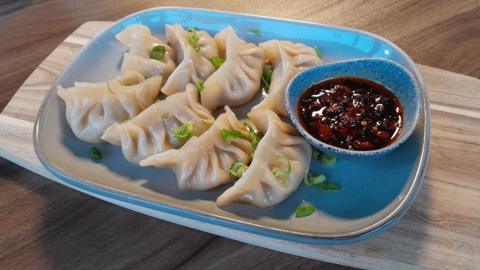 A plate of eight gyoza dumplings with a crimped edge to the pastry, dressed with green slices of spring onion, with a small dish of crispy chilli sauce on the side, all sitting on a wooden board.