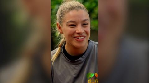 A woman smiling. Her blonde hair is tied back into a plait. She wears a grey T-shirt.