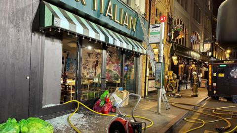 The front of Ask Italian on George Street. One of the windows is smashed with fire hoses going into it. A fire engine and two firefighters are on the street.