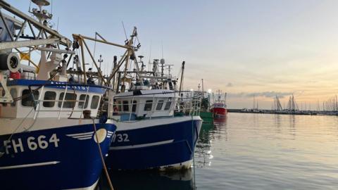 Fishing boats moored up in Brixham as the sun rises. The water is calm and the ski is turning pink. There are yachts tied up the other side of the harbour.