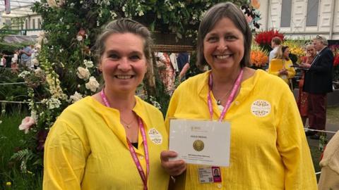 Two women stand next to each other, smiling at the camera. They are both wearing bright yellow blouses and pink lanyards. The woman on the left has brown and grey hair pulled back into a ponytail. The woman on the right has bobbed brown and grey hair and is holding an award with the words "RHS" and "GOLD MEDAL" on it.