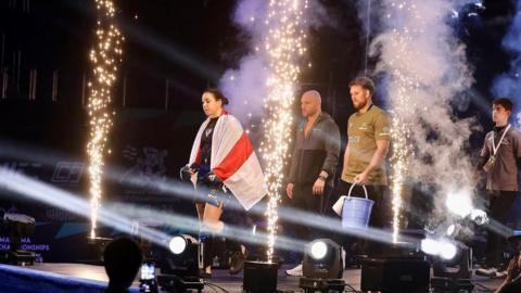 Alanna Pritchard walks across a stage with lights and fireworks. She is wearing black T-shirt and shorts and has a white and red flag draped over her shoulders, and shorts. Two men are standing behind her on the stage.