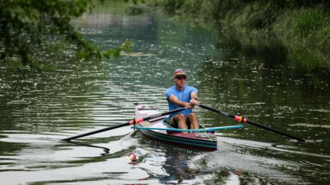 Mike Ellicock, wearing a blue t-shirt and shorts, rows his canoe on a stretch of open water.
