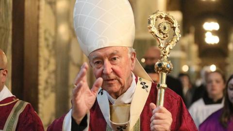 Cardinal Vincent Nichols holds up his hand while walking along. 