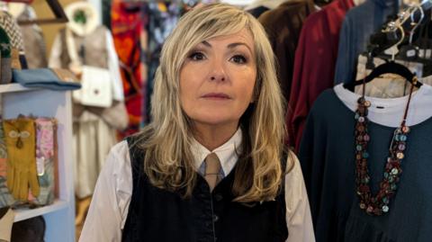 A woman standing looking at the camera with a blank look on her face. She is surrounded by clothes racks.