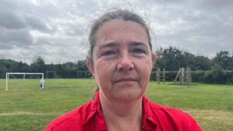 Ann Wilshaw has brown hair which is tied back. She is wearing a red shirt. Behind her is a green playing field with a white football goal post.