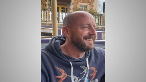 Tantalus St John-Ives smiling and looking to the right of the camera. He has a shaved head, moustache and beard and is wearing a navy hoodie with a pattern of swallows on it. The side of a building is in the background. 