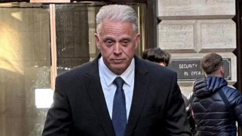 Ryan Cecil looking down as he walks outside the court. He has white hair and is wearing a dark suit, white shirt and blue tie.